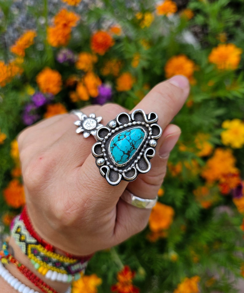 Op de afbeelding: Een zilveren ring met een turquoise steen in een decoratief ontwerp. De ring wordt gedragen op een vinger met meerdere armbanden.