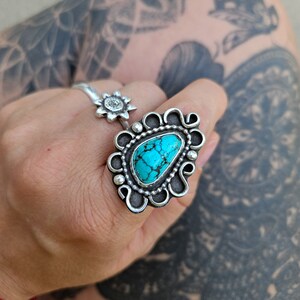 Op de afbeelding: Een zilveren ring met een turquoise steen in een decoratief ontwerp. De ring wordt gedragen op een hand met een tatoeage.