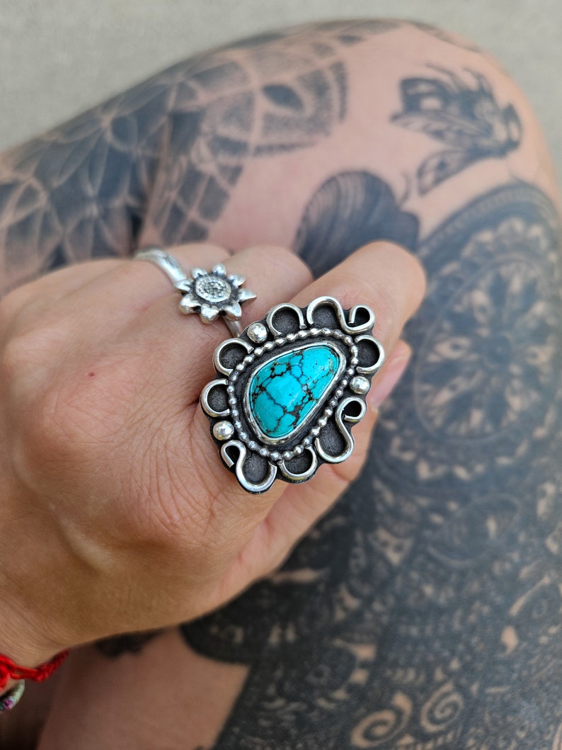 Op de afbeelding: Een zilveren ring met een turquoise steen in een decoratief ontwerp. De ring wordt gedragen op een hand met een tatoeage.
