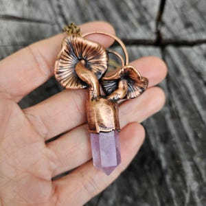 Puede incluir: Un colgante de cobre en forma de hongo con una punta de cristal de amatista púrpura. El colgante está en una cadena y se sostiene en una mano.