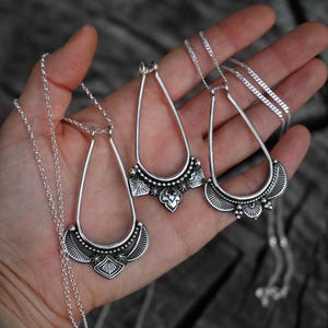 K&ouml;nnte beinhalten: Drei silberfarbene Anh&auml;ngerhalsketten mit tropfenf&ouml;rmigen Rahmen und detaillierten Designs. Die Halsketten werden in einer Hand gehalten, was die Handwerkskunst zeigt. Die Anh&auml;nger haben komplizierte Muster und sind an filigranen Ketten aufgeh&auml;ngt.