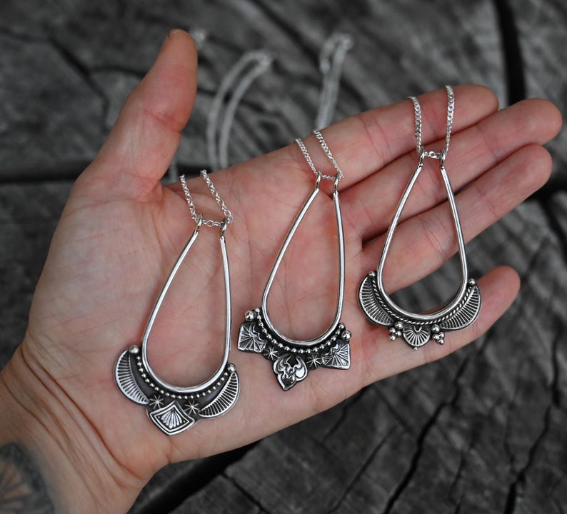K&ouml;nnte beinhalten: Drei silberfarbene Anh&auml;ngerhalsketten mit tropfenf&ouml;rmigen Rahmen und verzierten unteren Abschnitten. Die Anh&auml;nger werden auf einer Hand vor einem dunklen, strukturierten Hintergrund pr&auml;sentiert. Die Halsketten haben filigrane Kettenglieder.