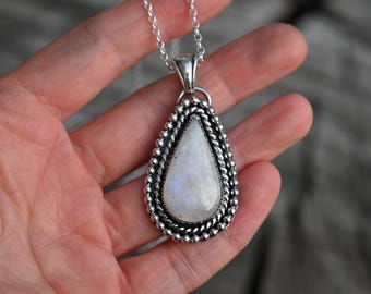 May include: A silver pendant necklace featuring a pear-shaped moonstone. The moonstone has a milky white and iridescent appearance, set within a beaded silver frame. The pendant hangs from a delicate silver chain.