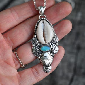 May include: A silver pendant necklace featuring a cowrie shell, an oval turquoise stone, and a small seashell. The pendant has intricate silver detailing and hangs from a delicate chain. A unique piece of jewelry.