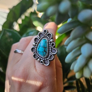 Op de afbeelding: Een zilveren ring met een turquoise steen in een decoratief ontwerp. De ring zit aan een vinger.