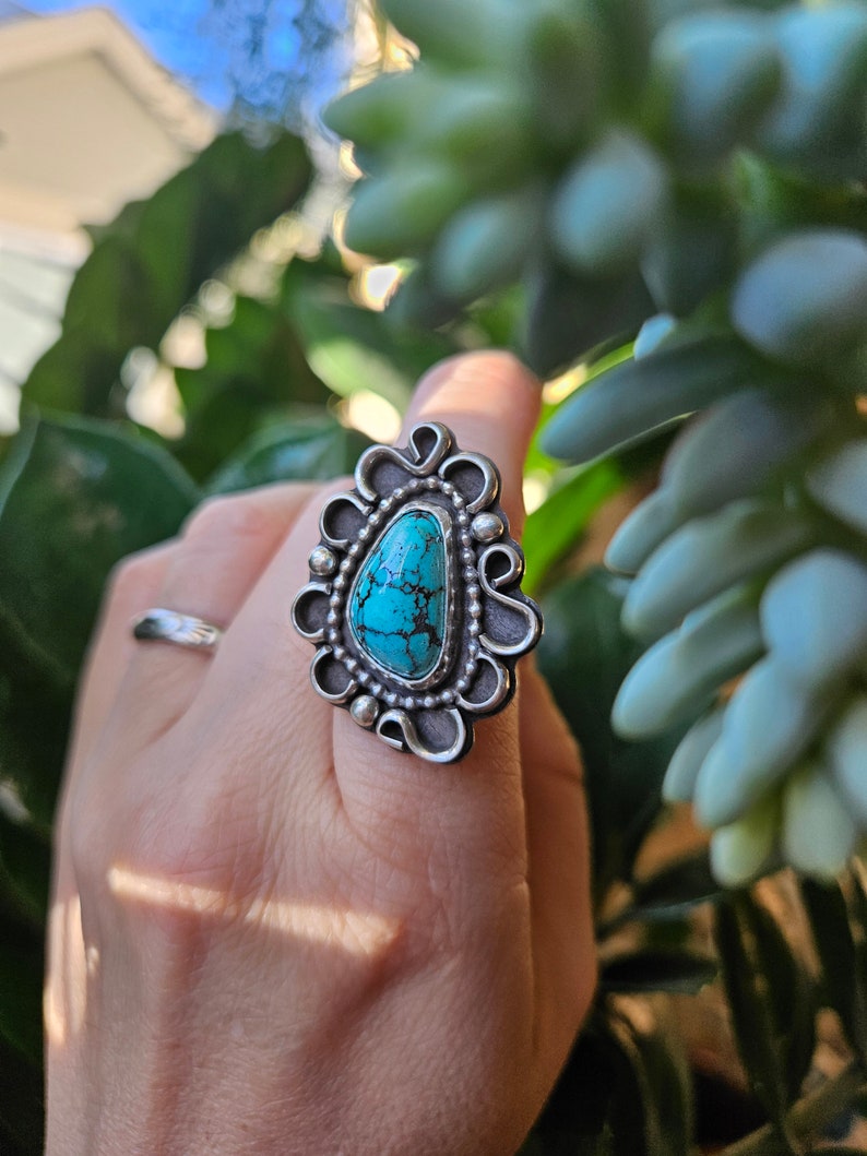 Op de afbeelding: Een zilveren ring met een turquoise steen in een decoratief ontwerp. De ring zit aan een vinger.