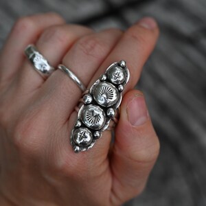 May include: A silver statement ring with a long, oval design featuring five textured, circular embellishments. The ring has a hammered metal finish and is worn on a finger. Two other silver rings are also visible.