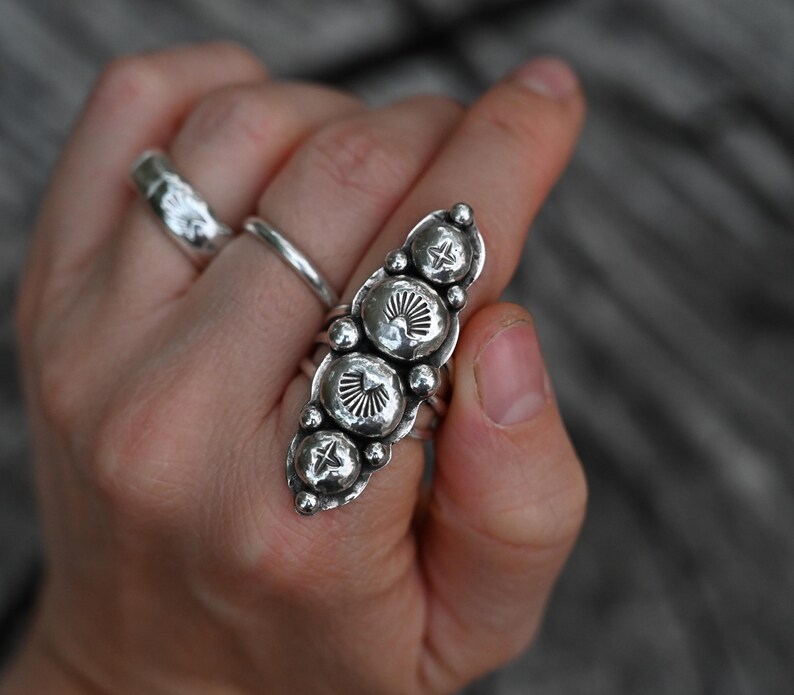 May include: A silver statement ring with a long, oval design featuring five textured, circular embellishments. The ring has a hammered metal finish and is worn on a finger. Two other silver rings are also visible.