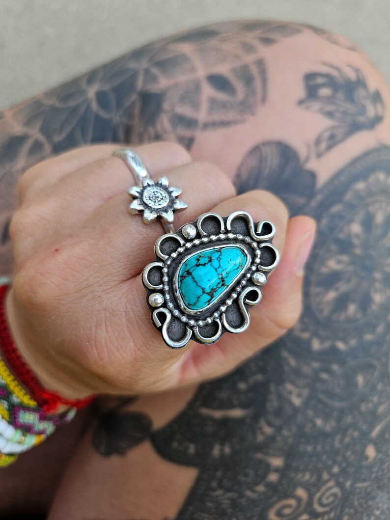 Op de afbeelding: Een zilveren ring met een turquoise steen in een decoratief ontwerp. De ring wordt gedragen op een vinger met een zilveren zonnebloemring op een andere vinger.