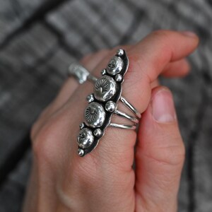 May include: A silver ring with a long, ornate design featuring multiple circular and oval-shaped elements. The ring has a detailed, textured surface and is worn on a finger. The ring's design includes small, raised details.