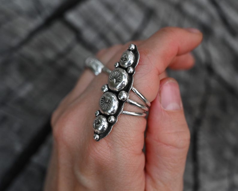 May include: A silver ring with a long, ornate design featuring multiple circular and oval-shaped elements. The ring has a detailed, textured surface and is worn on a finger. The ring's design includes small, raised details.