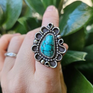 Op de afbeelding: Een zilveren ring met een turquoise steen in een decoratief ontwerp. De ring zit aan een vinger.