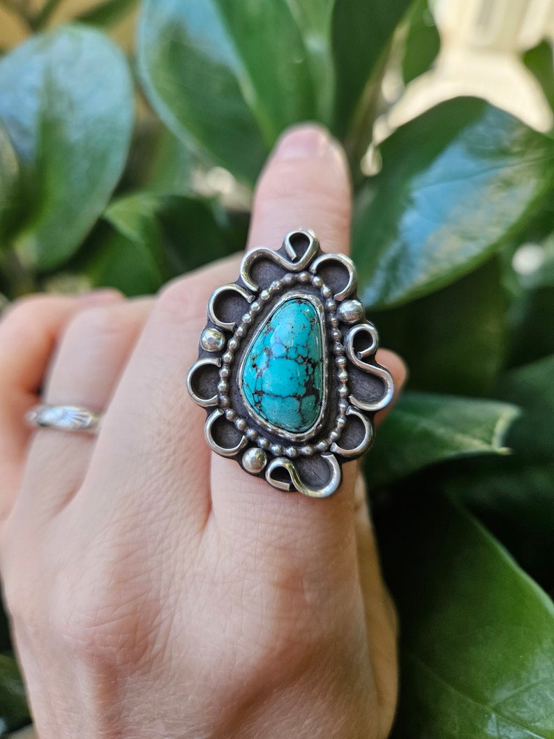 Op de afbeelding: Een zilveren ring met een turquoise steen in een decoratief ontwerp. De ring zit aan een vinger.