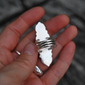 May include: A silver ring with a long, white, leaf-shaped stone. The ring's band is made of multiple coiled silver wires. The ring is held in a hand, with a blurred wooden background.