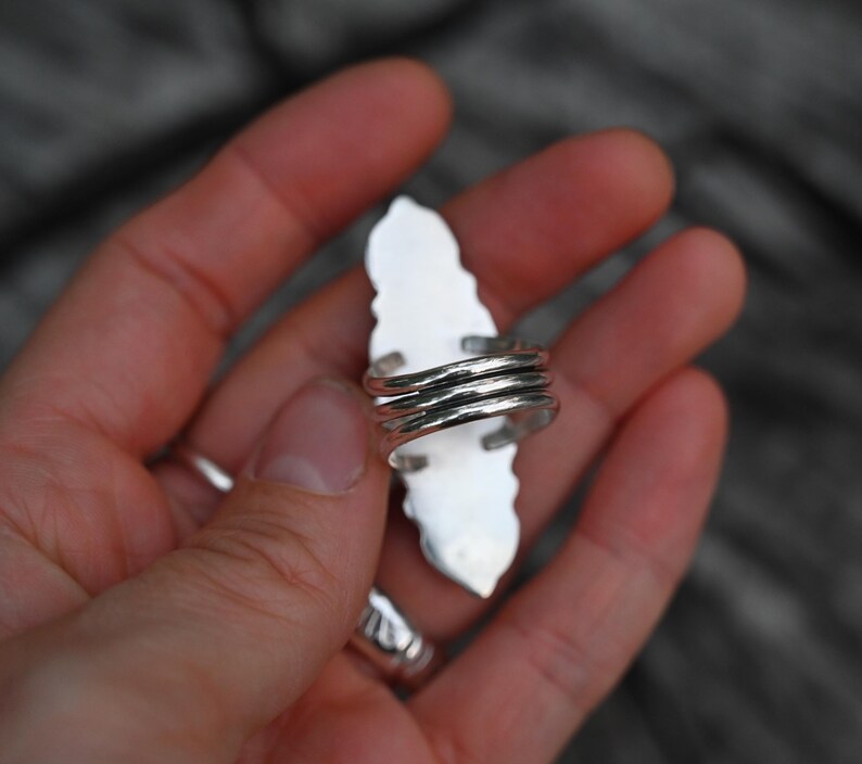 May include: A silver ring with a long, white, leaf-shaped stone. The ring's band is made of multiple coiled silver wires. The ring is held in a hand, with a blurred wooden background.