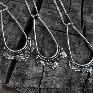 K&ouml;nnte beinhalten: Drei silberne Anh&auml;nger-Halsketten mit aufwendigen, detaillierten Designs. Jede Halskette hat einen tropfenf&ouml;rmigen Rahmen mit einem dekorativen, stilisierten Halbmond an der Basis. Die Anh&auml;nger liegen auf einer verwitterten, dunklen Holzoberfl&auml;che.