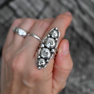 May include: A silver ring with a vertical design featuring four large, textured, circular elements. Each circular element has a sunburst pattern and is accented with small star-shaped details. Additional silver rings are also visible.