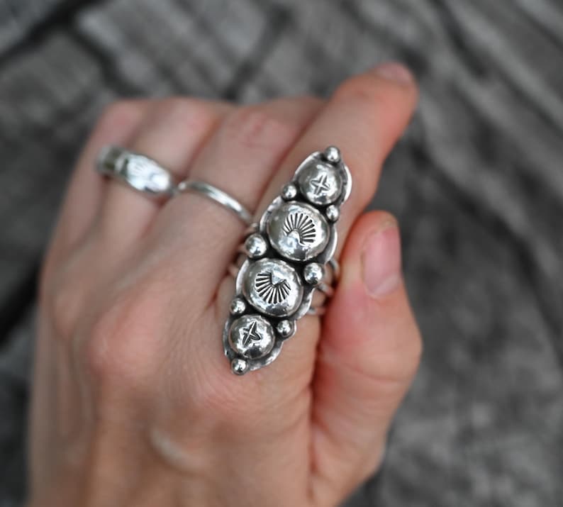 May include: A silver ring with a vertical design featuring four large, textured, circular elements. Each circular element has a sunburst pattern and is accented with small star-shaped details. Additional silver rings are also visible.