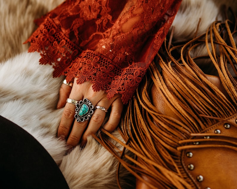 Op de afbeelding: Een vrouwenhand met meerdere ringen, waaronder een grote turquoise ring met een zilveren zetting. De hand rust op een bruine leren tas met franjes.