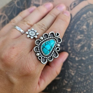 Op de afbeelding: Een zilveren ring met een turquoise steen in een decoratief ontwerp. De ring zit aan een vinger.
