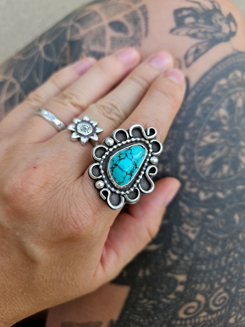 Op de afbeelding: Een zilveren ring met een turquoise steen in een decoratief ontwerp. De ring zit aan een vinger.