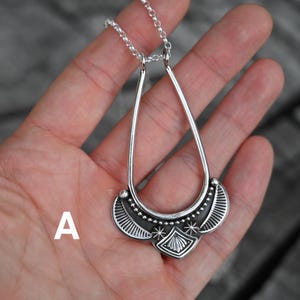 K&ouml;nnte beinhalten: Eine silberfarbene Anh&auml;ngerkette mit einem tropfenf&ouml;rmigen Design. Der Anh&auml;nger hat ein detailliertes Muster mit einer zentralen Diamantenform und Sternakzenten. Die Halskette wird in einer Hand vor einem verschwommenen Hintergrund gehalten.