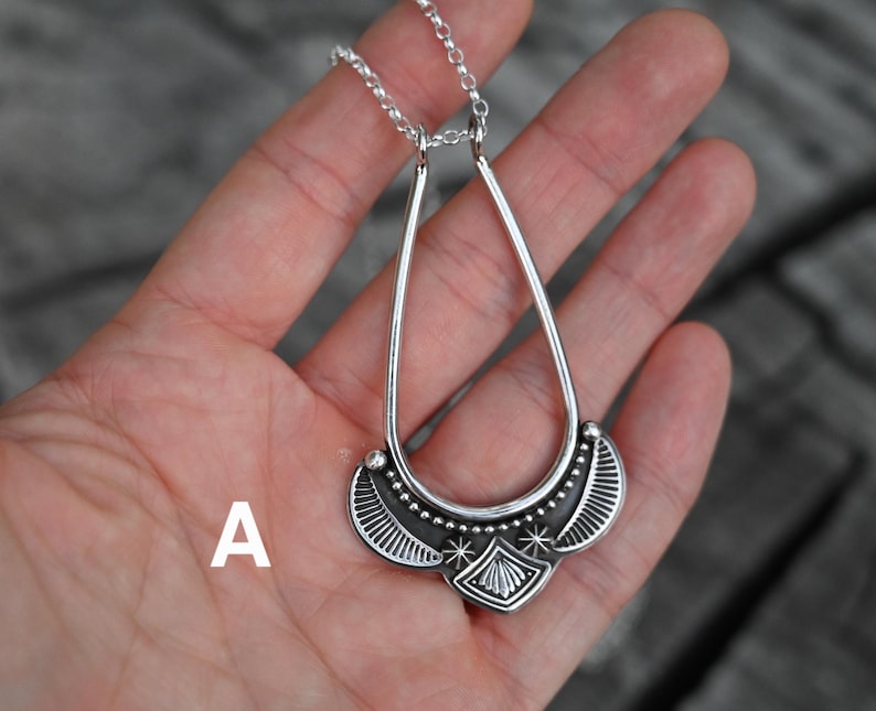 K&ouml;nnte beinhalten: Eine silberfarbene Anh&auml;ngerkette mit einem tropfenf&ouml;rmigen Design. Der Anh&auml;nger hat ein detailliertes Muster mit einer zentralen Diamantenform und Sternakzenten. Die Halskette wird in einer Hand vor einem verschwommenen Hintergrund gehalten.