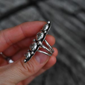 May include: A silver ring featuring a row of five textured, circular designs. The ring has a multi-band shank and small silver beads along the edges of the design. The ring is held in a hand against a blurred background.