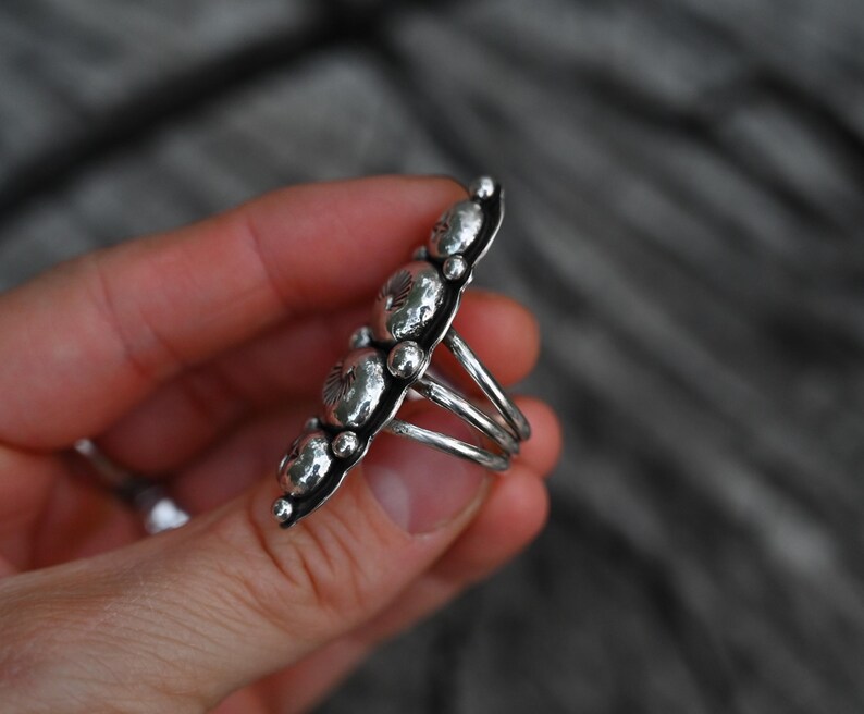 May include: A silver ring featuring a row of five textured, circular designs. The ring has a multi-band shank and small silver beads along the edges of the design. The ring is held in a hand against a blurred background.