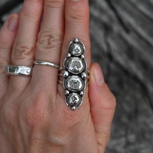 May include: A silver ring with an elongated design featuring four large, textured, circular elements. Each circular element has a sunburst pattern. The ring is worn on a finger, with two other silver rings visible.