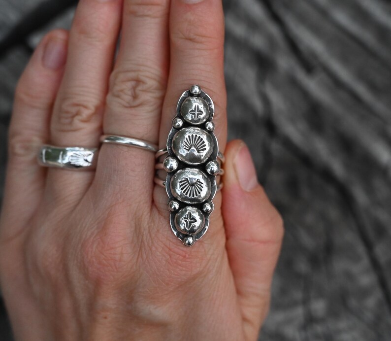 May include: A silver ring with an elongated design featuring four large, textured, circular elements. Each circular element has a sunburst pattern. The ring is worn on a finger, with two other silver rings visible.
