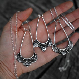 K&ouml;nnte beinhalten: Drei silberfarbene Anh&auml;ngerhalsketten mit tropfenf&ouml;rmigen Rahmen und detaillierten unteren Abschnitten. Die Halsketten werden auf einer Hand pr&auml;sentiert, wobei die zarte Kette und das verzierte Design der Anh&auml;nger gezeigt werden.