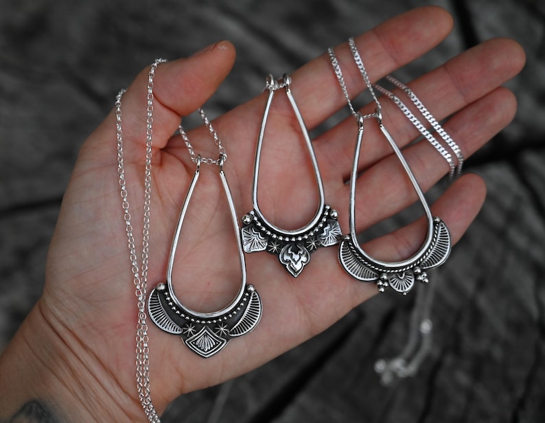 K&ouml;nnte beinhalten: Drei silberfarbene Anh&auml;ngerhalsketten mit tropfenf&ouml;rmigen Rahmen und detaillierten unteren Abschnitten. Die Halsketten werden auf einer Hand pr&auml;sentiert, wobei die zarte Kette und das verzierte Design der Anh&auml;nger gezeigt werden.
