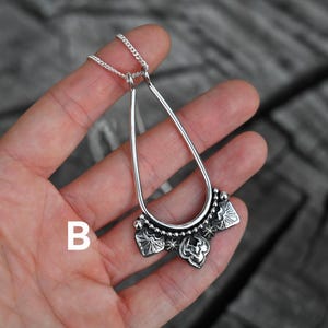 K&ouml;nnte beinhalten: Eine silberfarbene Anh&auml;ngerkette mit einem tropfenf&ouml;rmigen Rahmen und einem dekorativen unteren Bereich. Der untere Bereich weist florale und sternf&ouml;rmige Designs auf. Die Kette wird vor einem Holzgrund gezeigt.