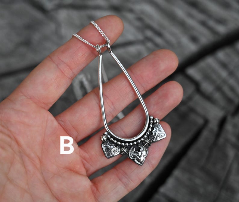K&ouml;nnte beinhalten: Eine silberfarbene Anh&auml;ngerkette mit einem tropfenf&ouml;rmigen Rahmen und einem dekorativen unteren Bereich. Der untere Bereich weist florale und sternf&ouml;rmige Designs auf. Die Kette wird vor einem Holzgrund gezeigt.