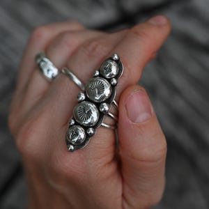 May include: A silver ring with a long, ornate design featuring five large, circular, textured embellishments. The ring has a series of smaller, round accents and multiple bands. Another silver ring is visible in the background.