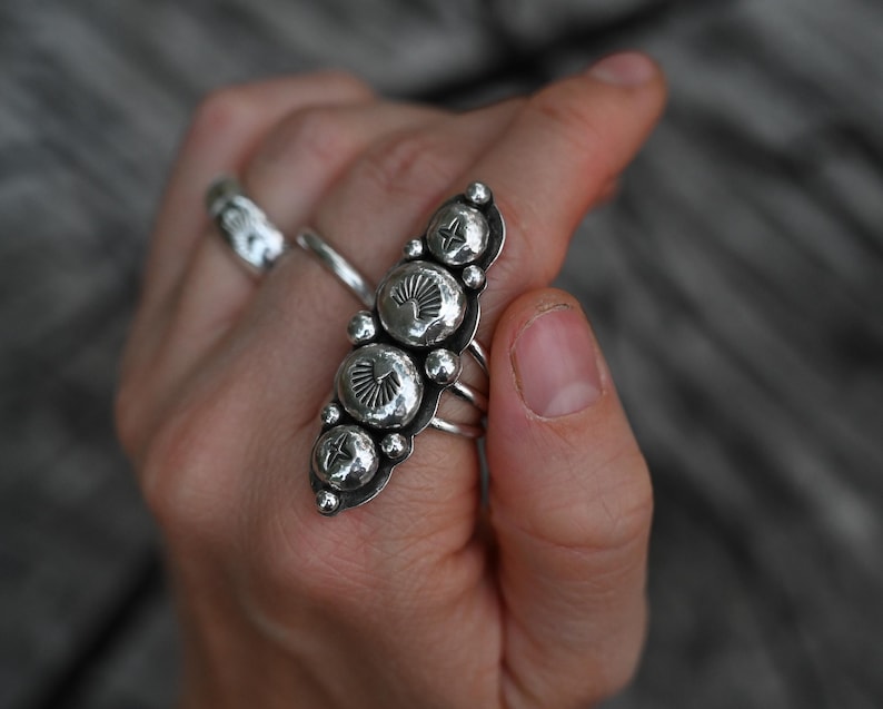 May include: A silver ring with a long, ornate design featuring five large, circular, textured embellishments. The ring has a series of smaller, round accents and multiple bands. Another silver ring is visible in the background.