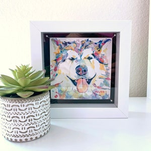 K&ouml;nnte beinhalten: Ein gerahmtes Aquarellbild eines bunten Hundes mit herausgestreckter Zunge. Der Hund befindet sich in einem wei&szlig;en Rahmen mit schwarzem Passepartout. Das Bild befindet sich auf einer wei&szlig;en Oberfl&auml;che mit einer kleinen Sukkulente in einem wei&szlig;en Topf.