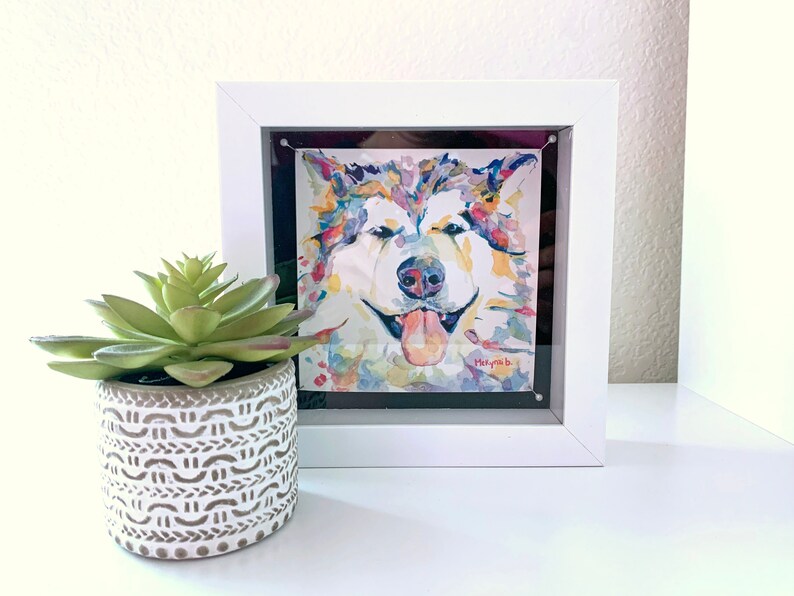 K&ouml;nnte beinhalten: Ein gerahmtes Aquarellbild eines bunten Hundes mit herausgestreckter Zunge. Der Hund befindet sich in einem wei&szlig;en Rahmen mit schwarzem Passepartout. Das Bild befindet sich auf einer wei&szlig;en Oberfl&auml;che mit einer kleinen Sukkulente in einem wei&szlig;en Topf.