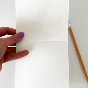 K&ouml;nnte beinhalten: Eine leere wei&szlig;e Karte mit leicht strukturierter Oberfl&auml;che. Die Karte ist in der Mitte gefaltet und wird von einer Hand gehalten.
