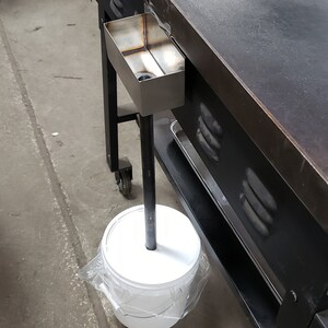 May include: A large, black metal griddle with a stainless steel grease trap. A white bucket is positioned below the griddle, connected by a black metal pole. The griddle is on wheels.