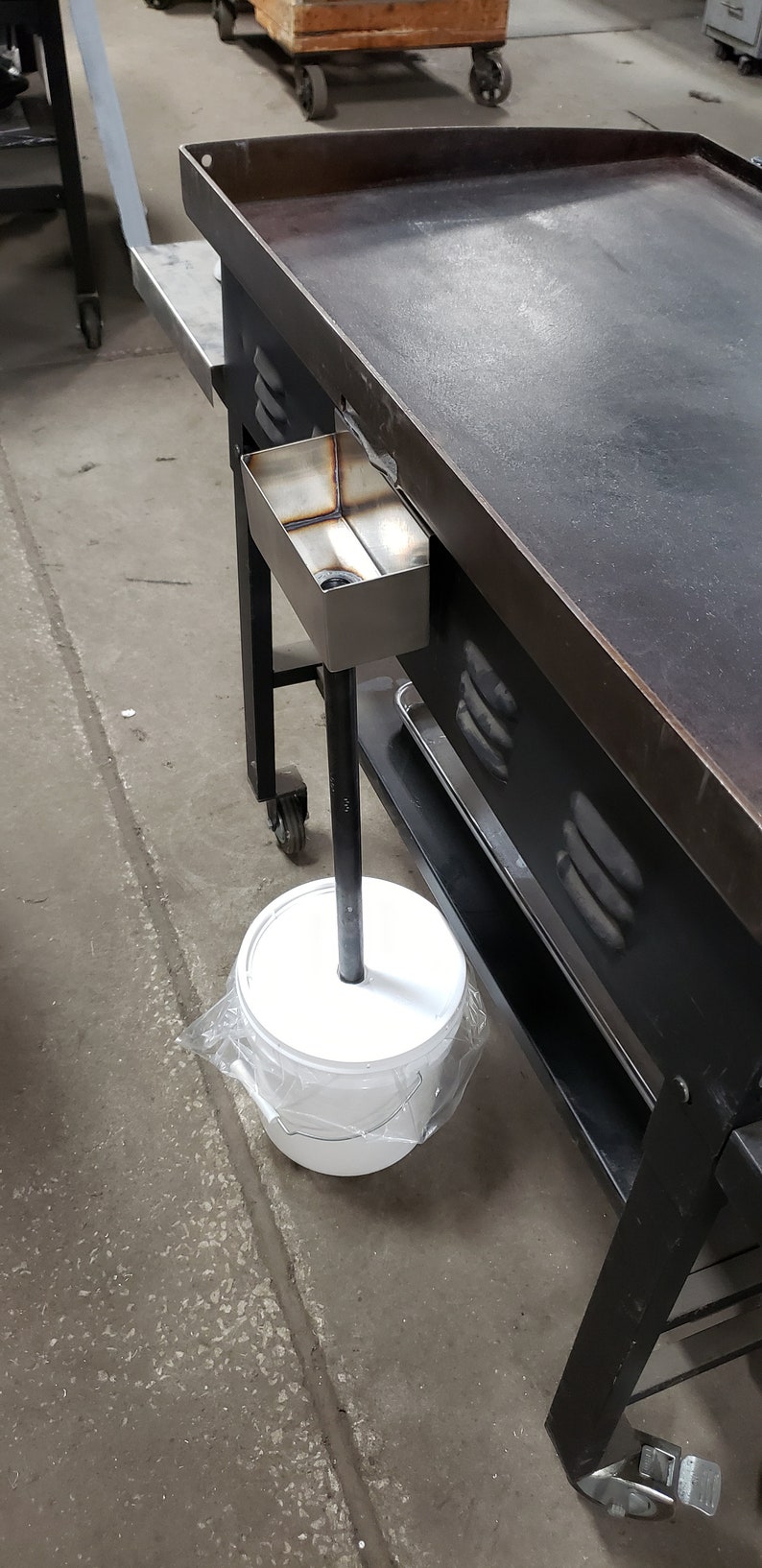 May include: A large, black metal griddle with a stainless steel grease trap. A white bucket is positioned below the griddle, connected by a black metal pole. The griddle is on wheels.