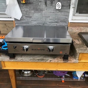 May include: A stainless steel Blackstone griddle with a black base and orange logo. The griddle is sitting on a wooden countertop with a granite surface. The griddle has two knobs and a side shelf.
