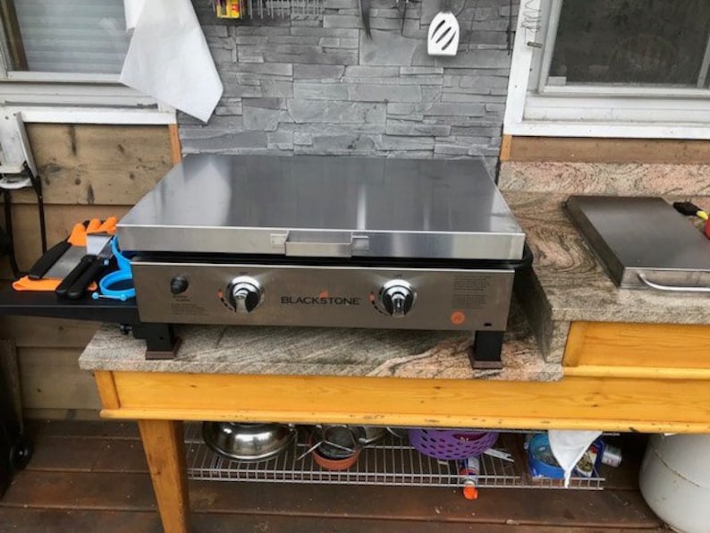 May include: A stainless steel Blackstone griddle with a black base and orange logo. The griddle is sitting on a wooden countertop with a granite surface. The griddle has two knobs and a side shelf.