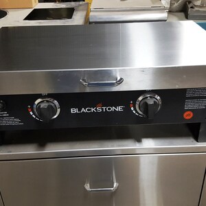 May include: Blackstone flat top grill with two knobs and a stainless steel top. The grill is on a stainless steel stand with a drawer.