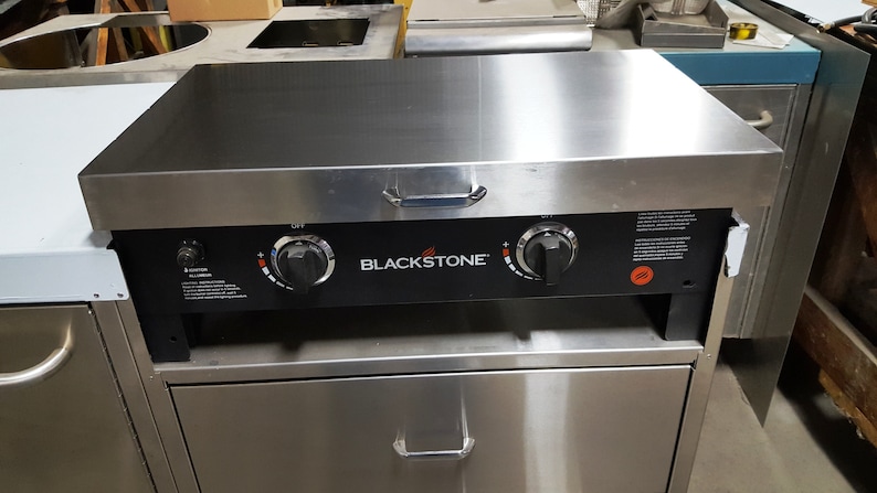 May include: Blackstone flat top grill with two knobs and a stainless steel top. The grill is on a stainless steel stand with a drawer.