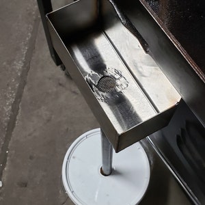 May include: A stainless steel rectangular tray with a mesh drain, mounted on a metal pole and a white circular base. The tray is positioned near a black, angled surface.