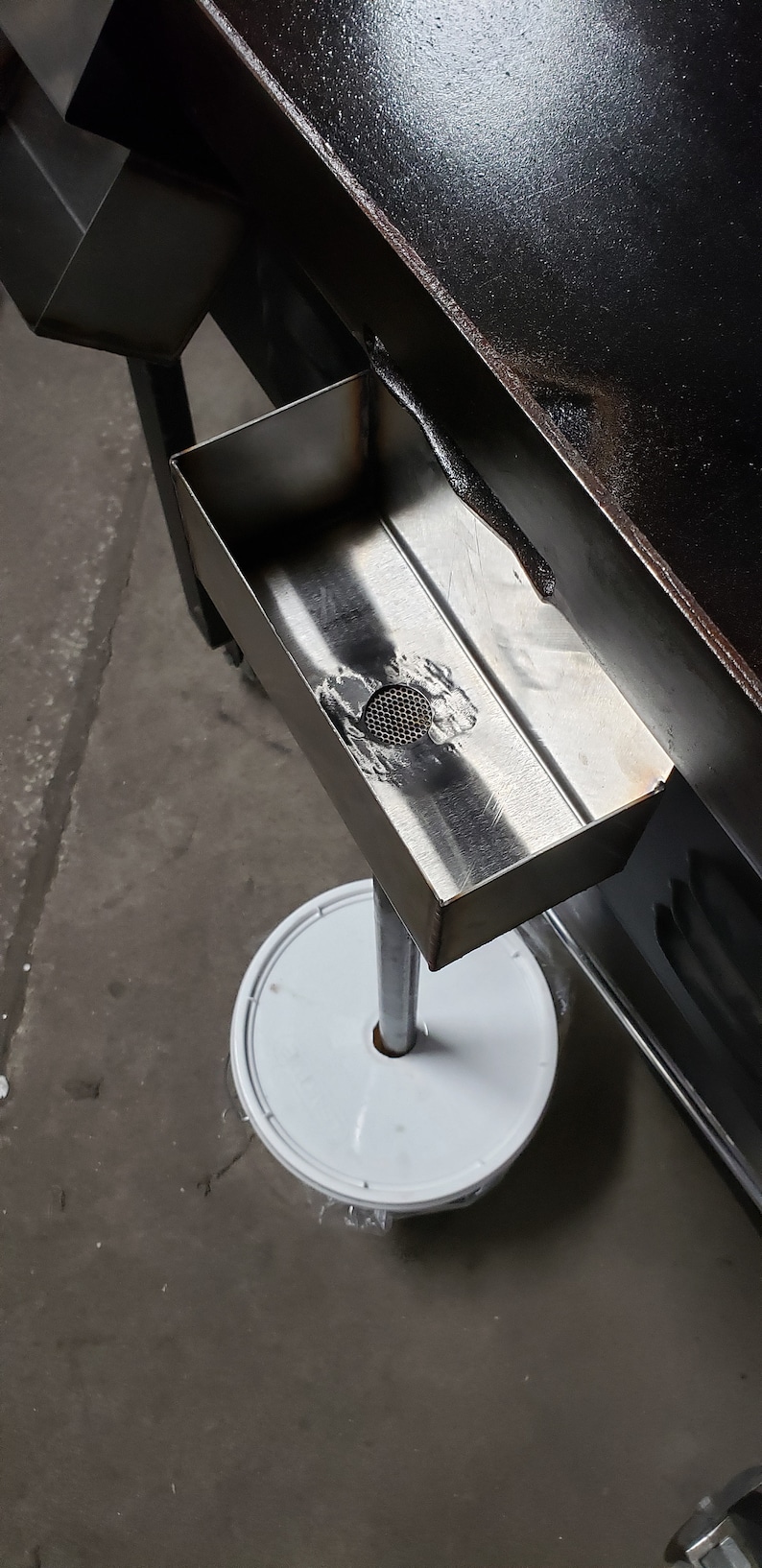 May include: A stainless steel rectangular tray with a mesh drain, mounted on a metal pole and a white circular base. The tray is positioned near a black, angled surface.