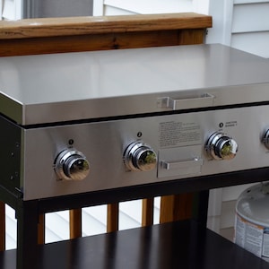 May include: A black and stainless steel gas grill with four burner knobs and a stainless steel top. The grill is on a black metal stand with a lower shelf.