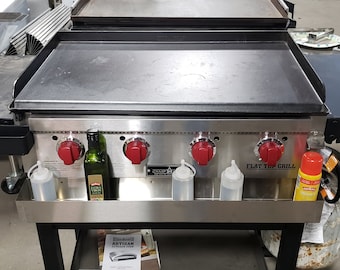May include: A stainless steel flat top grill with four red knobs and a black side shelf. The grill has a stainless steel shelf underneath with a bottle of olive oil, two squeeze bottles, and a can of spray oil.